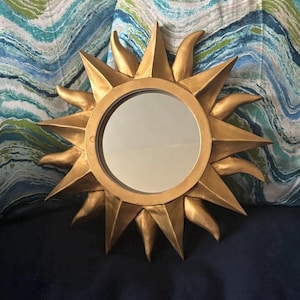 May include: A gold-colored sunburst mirror with a round mirrored center. The sunburst design features pointed rays and curved accents. The mirror is set against a dark blue surface and patterned pillows.