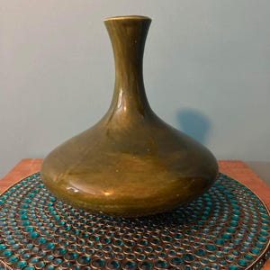 May include: A green, pear-shaped vase with a narrow neck. The vase sits on a round, decorative coaster with teal-colored jewels. The vase has a glossy finish and a dark green color.