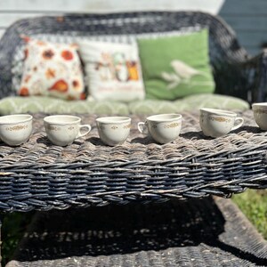 May include: Six off-white teacups with gold floral detailing are arranged on a dark wicker table. The cups have small handles and a scalloped rim. Pillows with fall-themed patterns rest on a wicker bench in the background.