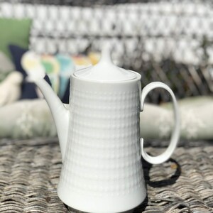 May include: A white ceramic teapot with a textured, dotted pattern. The teapot has a curved handle, a spout, and a lid with a small knob. The teapot is sitting on a woven surface, with a blurred background of outdoor furniture.