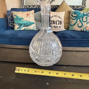 May include: A clear, cut-glass decanter with a bulbous base and a tall, flared neck. The decanter features vertical and diamond-shaped cut patterns. A measuring tape shows the decanter is approximately 9 inches tall. The background includes a blue couch and decorative pillows.