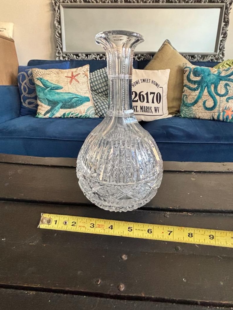 May include: A clear, cut-glass decanter with a bulbous base and a tall, flared neck. The decanter features vertical and diamond-shaped cut patterns. A measuring tape shows the decanter is approximately 9 inches tall. The background includes a blue couch and decorative pillows.