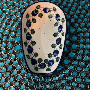 May include: White ceramic spoon rest with a blue rim and a hand-painted design of blueberries and green leaves. The spoon rest is oval-shaped and sits on a blue jeweled surface.
