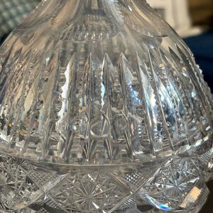 May include: A clear, cut-glass decanter with intricate geometric patterns. The upper portion features vertical cuts, while the lower section showcases a starburst design. The glass reflects light, creating a sparkling effect. The decanter is likely used for serving beverages.