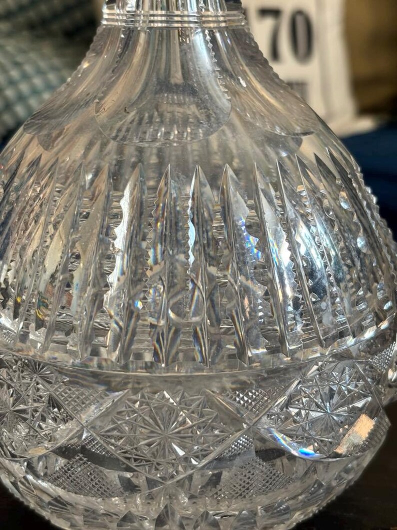 May include: A clear, cut-glass decanter with intricate geometric patterns. The upper portion features vertical cuts, while the lower section showcases a starburst design. The glass reflects light, creating a sparkling effect. The decanter is likely used for serving beverages.