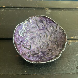 May include: A small, round, hand-formed ceramic dish featuring a deep purple glaze with a metallic sheen. The surface is textured with a repeating pattern of raised, irregular, swirling shapes and dark, recessed grooves throughout the interior.