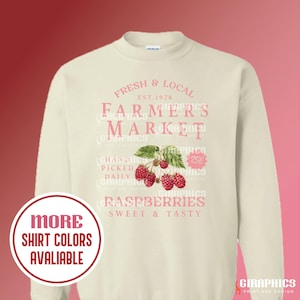 May include: A cream colored sweatshirt with a pink graphic design that says "Fresh & Local Est. 1978 Farmers Market Hand Picked Daily Raspberries Sweet & Tasty". The graphic also features a cluster of red raspberries.