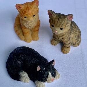 May include: Three small ceramic cat figurines. One is orange and white, one is brown and white, and one is black and white. The black and white cat is lying down, while the other two are sitting up.
