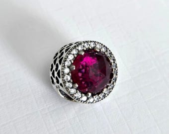 May include: A silver charm with a large, round, dark pink gemstone surrounded by a halo of smaller, clear gemstones. The charm has a delicate, patterned design.
