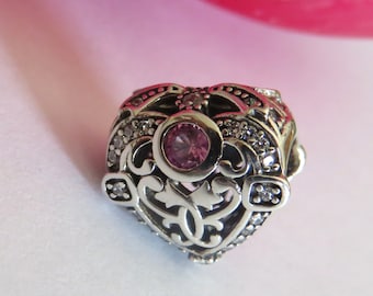 May include: A heart-shaped charm with a central pink gemstone and intricate filigree detailing. The charm is silver-toned and adorned with small, clear crystals. The design is ornate and elegant, suitable for jewelry.