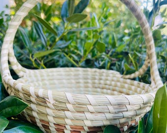 May include: A round woven basket featuring a high, arched handle. The basket is constructed from light beige natural fibers with a single horizontal band of reddish-brown material woven into the lower section of the circular body.