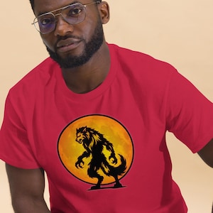 May include: A red short-sleeved t-shirt featuring a circular graphic on the chest. The graphic depicts a black silhouette of a werewolf standing in front of a large, textured orange full moon. The shirt has a crew neckline and a standard fit.