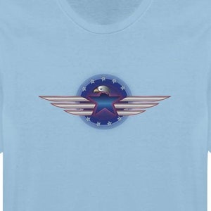 May include: Light blue t-shirt featuring a graphic of an eagle with outstretched wings. The eagle is set within a blue circle with white stars and a red and blue star in the center. The wings are silver and white.