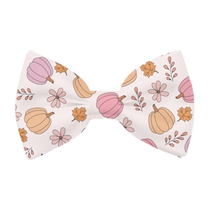 May include: A white bow tie with a repeating pattern of pumpkins, flowers, and leaves in shades of pink, orange, and brown. The bow tie is a classic shape, perfect for fall or autumn-themed events.