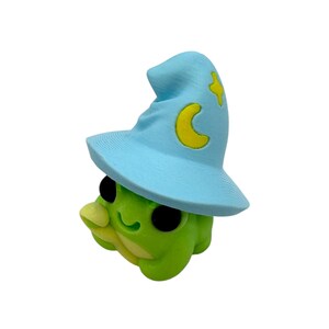 May include: A whimsical green frog figurine wearing a light blue wizard hat with a yellow crescent moon and star. The frog has large black eyes and a smiling expression, perfect for fantasy-themed decor.