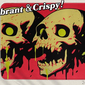 May include: A rectangular mat featuring a graphic design of two skulls with yellow eyes and teeth, set against a red background. The skulls have black outlines and dripping green accents. The text "Vibrant & Crispy!" is at the top. The mat measures 24" x 14".