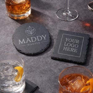 May include: A collection of slate coasters with drinks. One round coaster features the text "MADDY MARKED" and a leaf design. A square coaster displays "YOUR LOGO HERE". Other drinks include cocktails and a glass of water with a lemon peel.