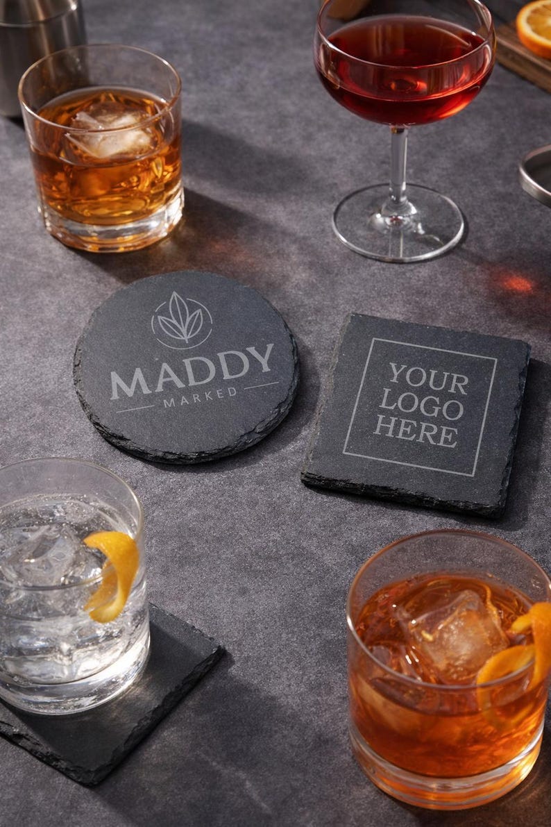 May include: A collection of slate coasters with drinks. One round coaster features the text "MADDY MARKED" and a leaf design. A square coaster displays "YOUR LOGO HERE". Other drinks include cocktails and a glass of water with a lemon peel.