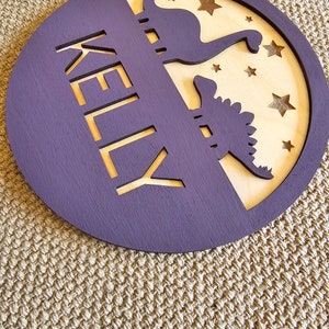 May include: A purple wooden circle with the name "KELLY" cut out in the center. The circle has a wood veneer insert with two dinosaur silhouettes and stars cut out.