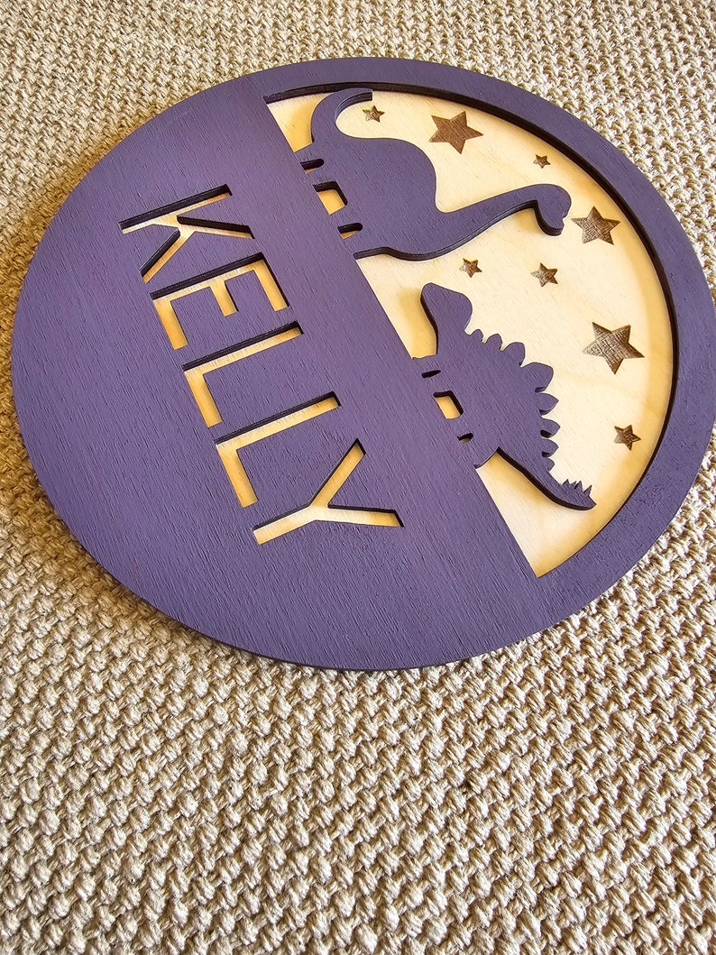 May include: A purple wooden circle with the name "KELLY" cut out in the center. The circle has a wood veneer insert with two dinosaur silhouettes and stars cut out.