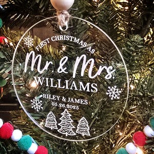 May include: Clear, circular Christmas ornament with the text "First Christmas as Mr. & Mrs. Williams" and the names "Riley & James" with the date "05.26.2025". The ornament has a snowflake and Christmas tree design. It hangs from a jute string and wooden bead.