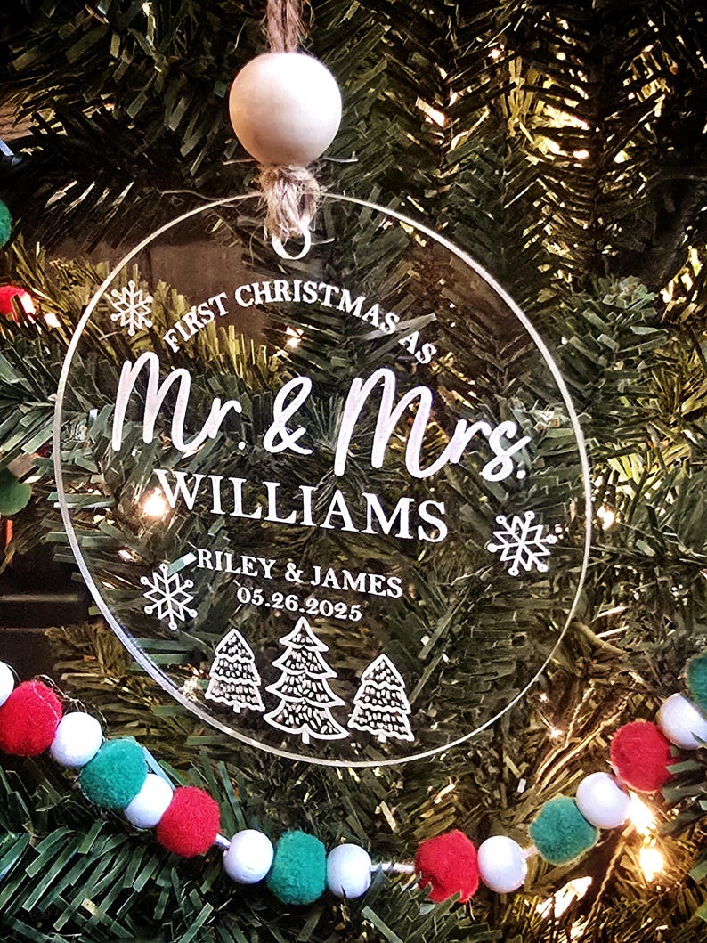 May include: Clear, circular Christmas ornament with the text "First Christmas as Mr. & Mrs. Williams" and the names "Riley & James" with the date "05.26.2025". The ornament has a snowflake and Christmas tree design. It hangs from a jute string and wooden bead.