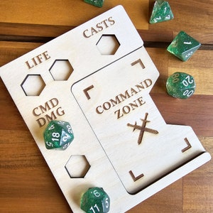May include: A wooden dice tray with compartments for different types of dice. The tray has labels for "Life", "CMD DMG", "CASTS", and "COMMAND ZONE". There are several green dice with white numbers on the tray.