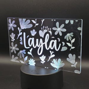 May include: A personalized LED night light with the name "Layla" in white script. The clear acrylic panel features a floral design in shades of blue and silver. The light sits on a black circular base.