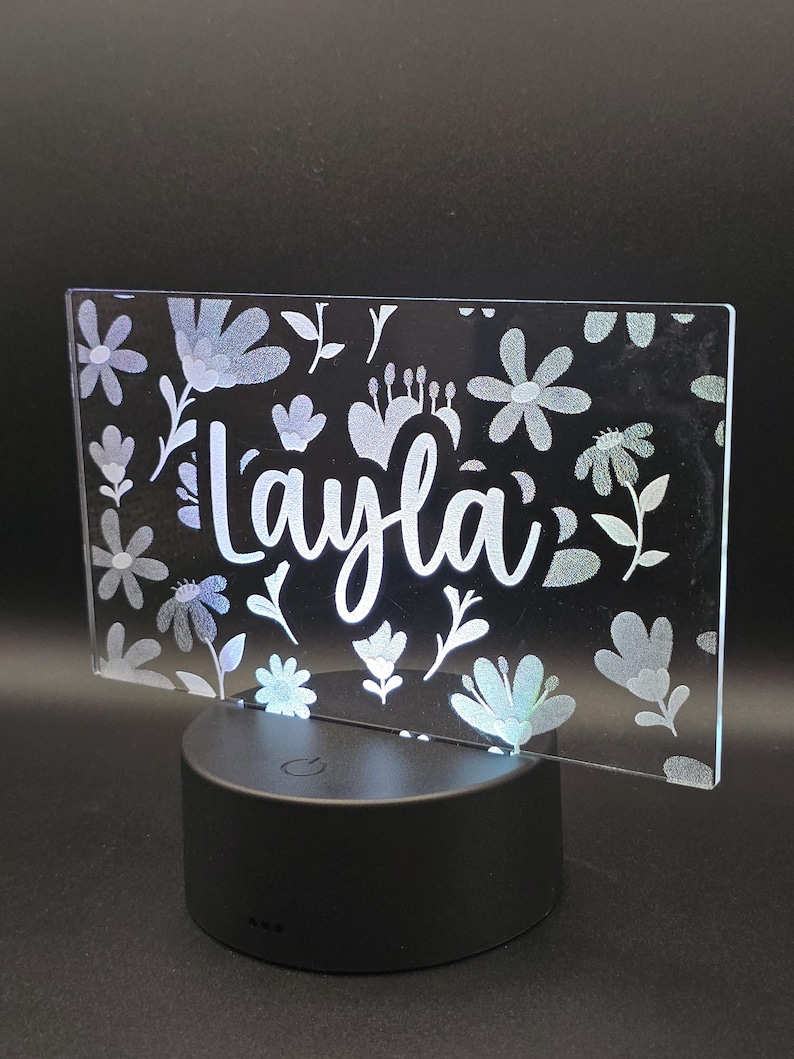 May include: A personalized LED night light with the name "Layla" in white script. The clear acrylic panel features a floral design in shades of blue and silver. The light sits on a black circular base.