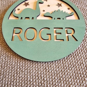 May include: A green wooden circle with a cutout of the name "ROGER" and two dinosaur silhouettes. The background is a light wood with stars.