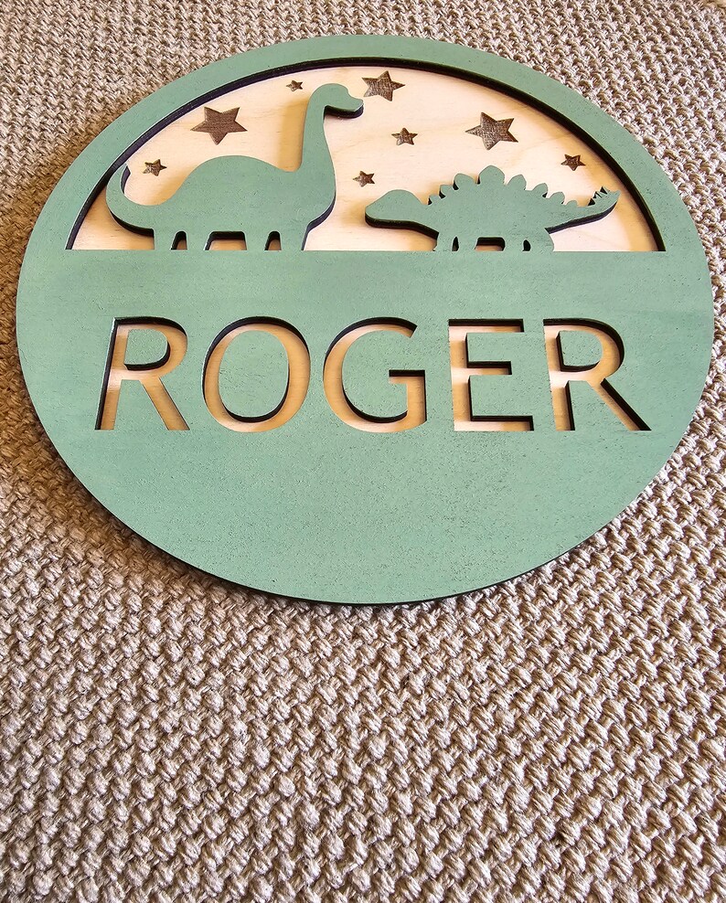 May include: A green wooden circle with a cutout of the name "ROGER" and two dinosaur silhouettes. The background is a light wood with stars.