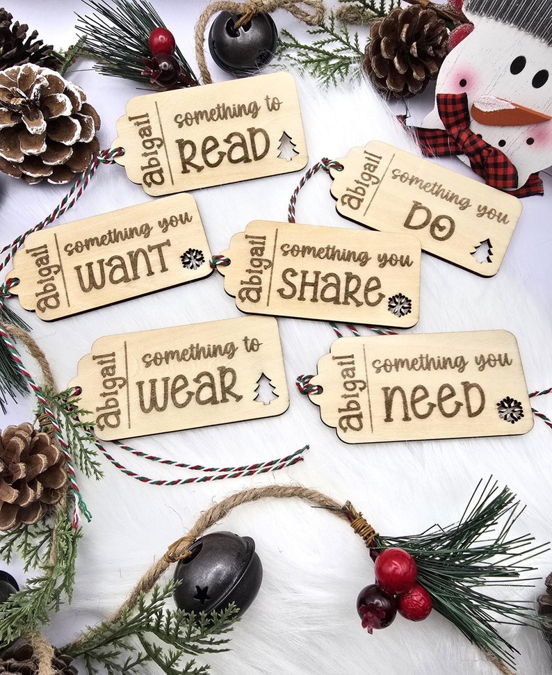 Pu&ograve; includere: Cinque etichette regalo in legno con finitura marrone rustica. Ogni etichetta &egrave; dotata di uno spago bianco e presenta il testo "abigail" e una frase diversa a tema natalizio. Le frasi sono "qualcosa da leggere", "qualcosa che vuoi", "qualcosa da condividere", "qualcosa da indossare" e "qualcosa di cui hai bisogno".