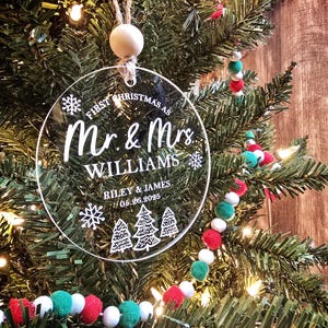 May include: A clear, circular Christmas ornament with the text "First Christmas as Mr. & Mrs. Williams" and the names Riley & James, with the date 05.26.2025. The ornament hangs from a wooden bead and twine, set against a Christmas tree.