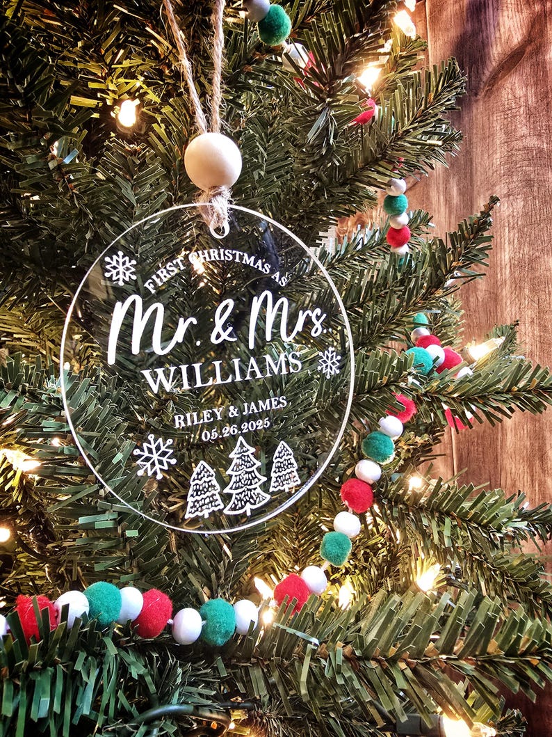 May include: A clear, circular Christmas ornament with the text "First Christmas as Mr. & Mrs. Williams" and the names Riley & James, with the date 05.26.2025. The ornament hangs from a wooden bead and twine, set against a Christmas tree.
