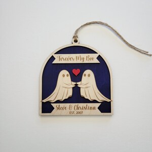 May include: Wooden ornament with two ghosts holding hands and a red heart. The ornament is painted blue and has the text "Forever My Boo" and "Steve & Christina EST. 2007".