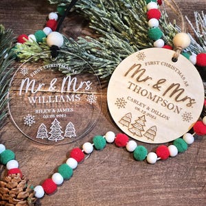 May include: Two Christmas ornaments with the text "First Christmas as Mr. & Mrs." One is clear acrylic, the other wood. The clear ornament says "Williams" and the wooden one says "Thompson". Both have names and dates. A garland with red, white, and green beads is in the image.