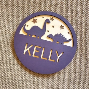 May include: Purple wooden circle with a cutout of two dinosaurs and stars. The name "KELLY" is cut out in the center.