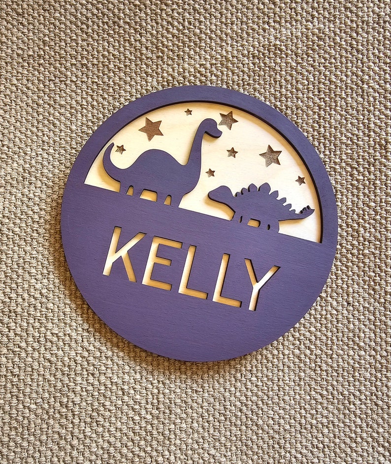 May include: Purple wooden circle with a cutout of two dinosaurs and stars. The name "KELLY" is cut out in the center.