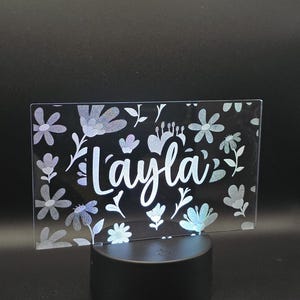 May include: A rectangular, illuminated sign with the name "Layla" in white script, surrounded by floral designs. The sign is supported by a black, circular base. The flowers are in shades of white and blue.
