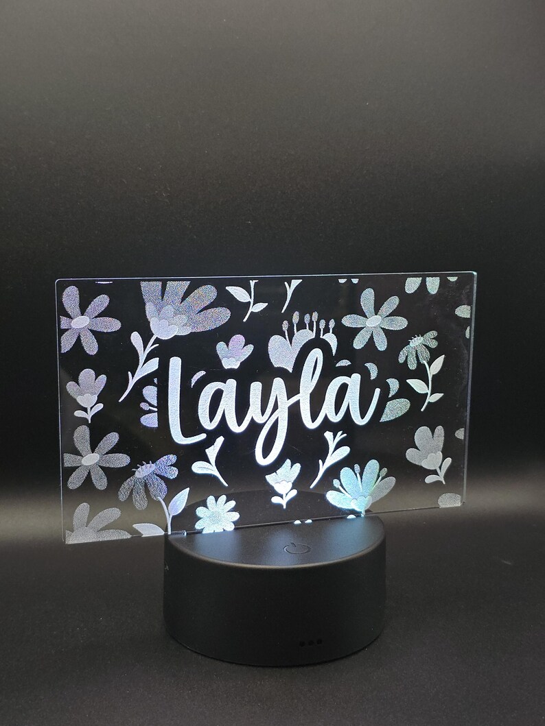 May include: A rectangular, illuminated sign with the name "Layla" in white script, surrounded by floral designs. The sign is supported by a black, circular base. The flowers are in shades of white and blue.
