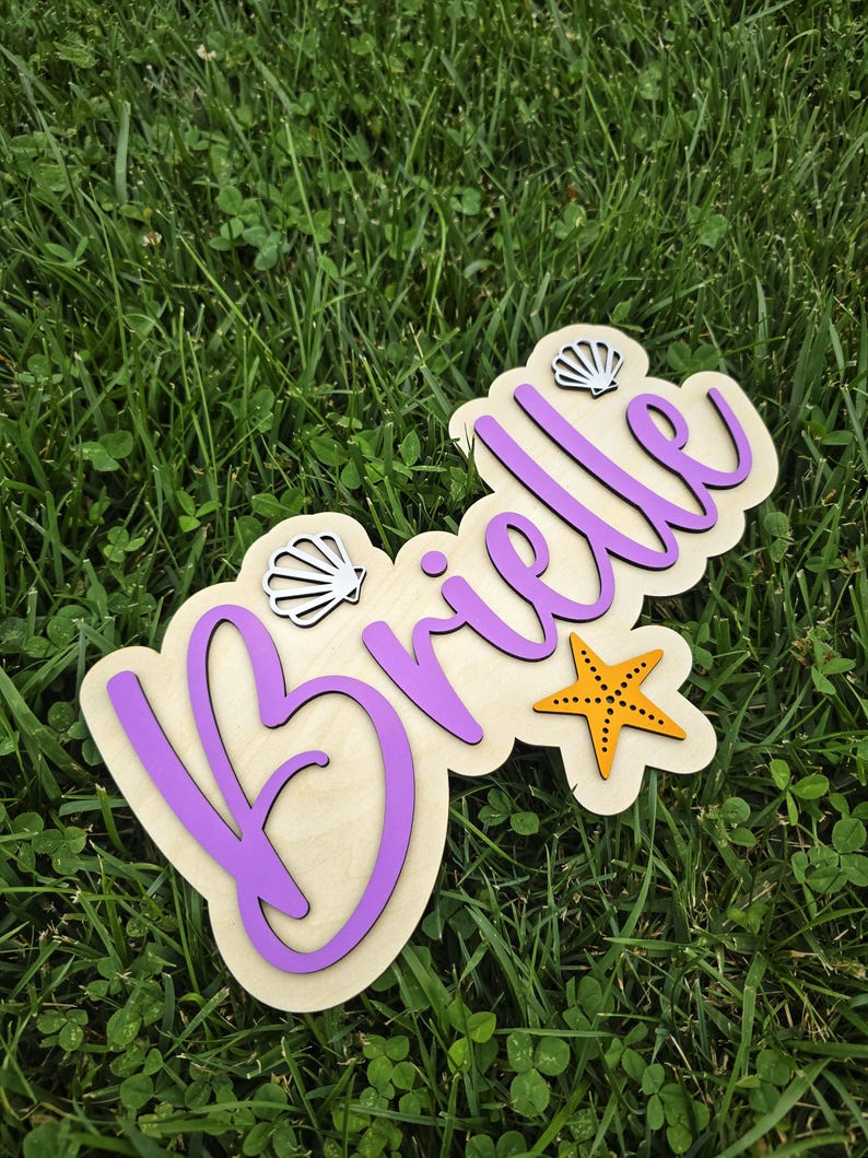 May include: Wooden sign with the name "Brielle" in purple script, accented with a starfish and two seashell cutouts. The sign is set against a background of green grass.