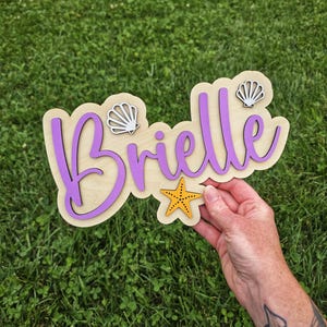 May include: A wooden sign with the name "Brielle" in purple script, layered over a light wood base. The sign is decorated with a yellow starfish and two seashell accents. The sign is held against a background of green grass.
