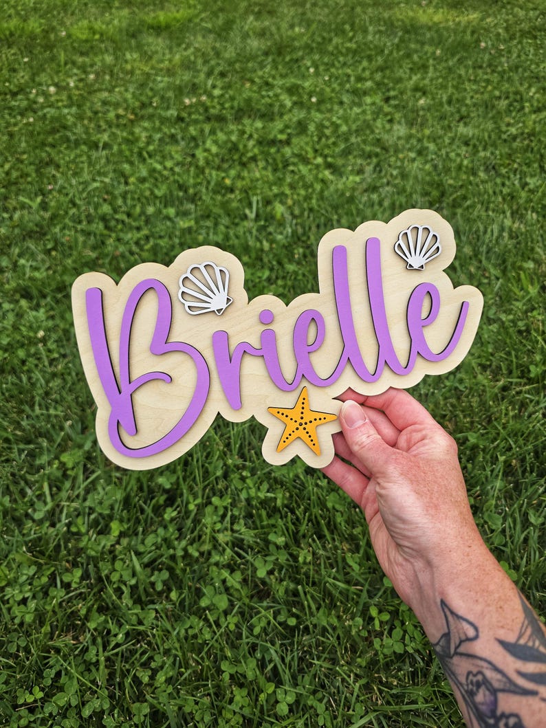 May include: A wooden sign with the name "Brielle" in purple script, layered over a light wood base. The sign is decorated with a yellow starfish and two seashell accents. The sign is held against a background of green grass.
