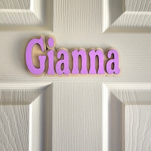 May include: A wooden name sign with the name "Gianna" painted in a light purple color. The sign is made of natural wood and has a rustic look.