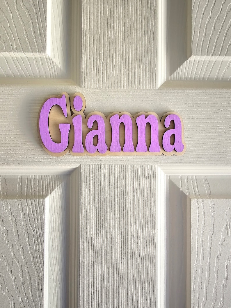 May include: A wooden name sign with the name "Gianna" painted in a light purple color. The sign is made of natural wood and has a rustic look.