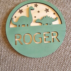 May include: A green wooden circle with a cutout of two dinosaurs and stars. The name "ROGER" is cut out in the center.