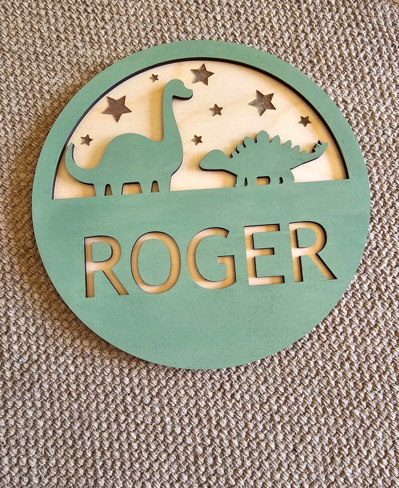 May include: A green wooden circle with a cutout of two dinosaurs and stars. The name "ROGER" is cut out in the center.