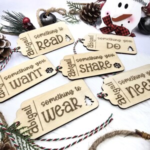 Pu&ograve; includere: Cinque etichette regalo in legno con un tema natalizio festoso. Ogni etichetta &egrave; decorata con una frase diversa: "qualcosa da leggere", "qualcosa che vuoi", "qualcosa da indossare", "qualcosa che fai" e "qualcosa che condividi".