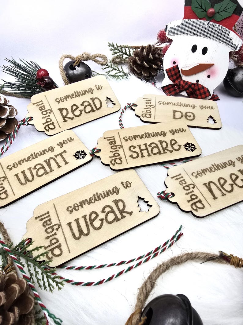 Pu&ograve; includere: Cinque etichette regalo in legno con un tema natalizio festoso. Ogni etichetta &egrave; decorata con una frase diversa: "qualcosa da leggere", "qualcosa che vuoi", "qualcosa da indossare", "qualcosa che fai" e "qualcosa che condividi".