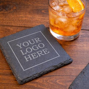May include: A square, dark gray slate coaster with the text "YOUR LOGO HERE" in a white, outlined font. A glass of amber-colored drink with ice and an orange slice sits nearby. The coaster is on a dark wood surface.